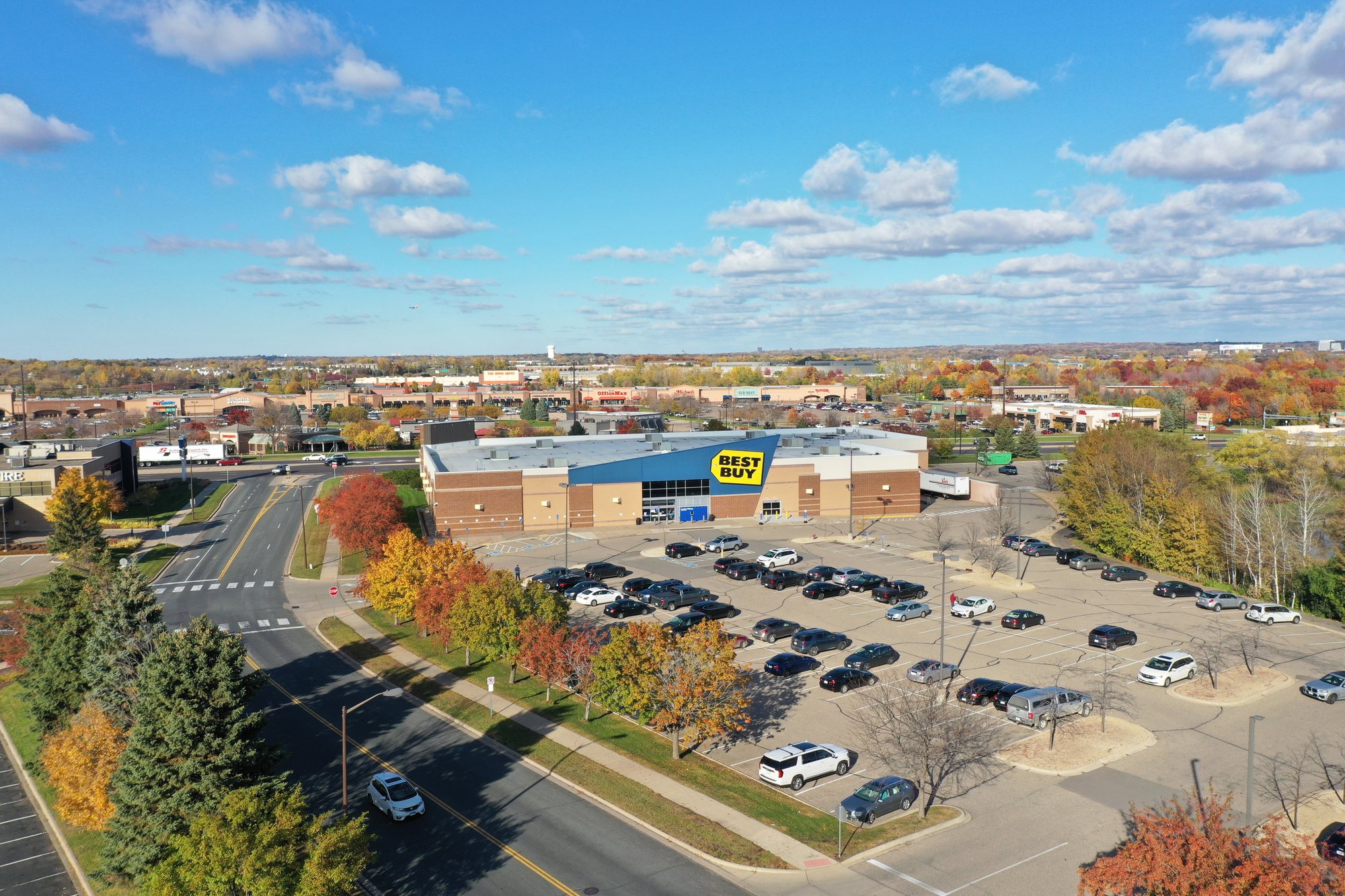Aerial view of a retail property, autumn trees, access roads, and the surrounding commercial district